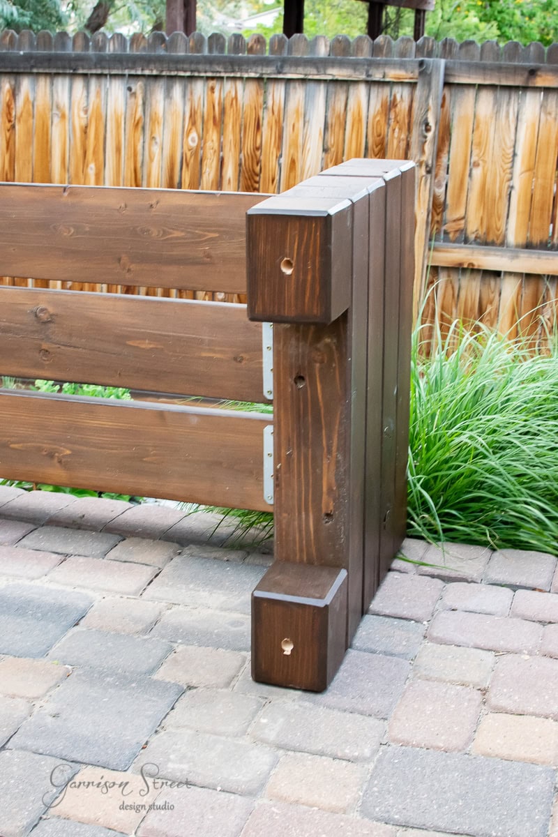 A wooden outdoor structure with vertical and horizontal beams is built on a stone patio, seamlessly integrating an outdoor sofa for comfort. It's designed for support or seating, with grass and a wooden fence in the background. The wood has a rich, dark finish.