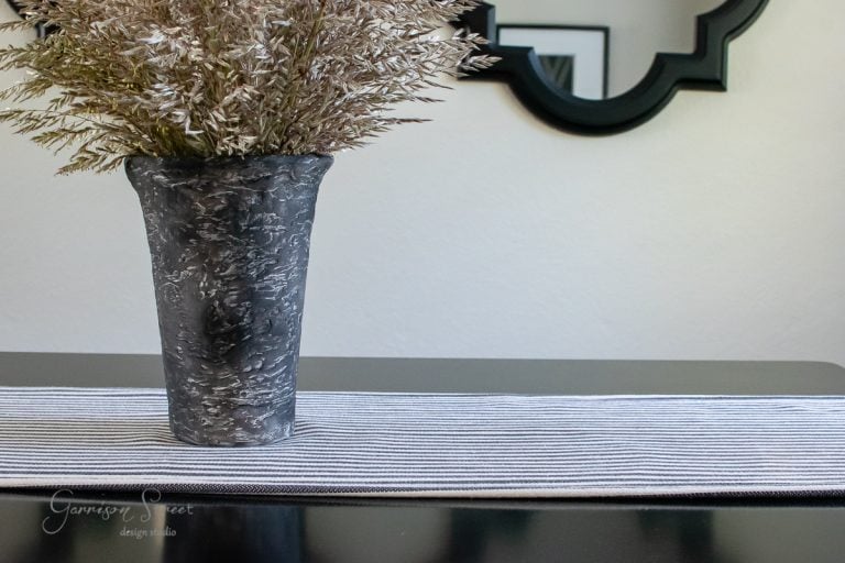 A textured artisan vase holding decorative grass sits on a black table with a striped runner. A black geometric mirror is visible in the background.