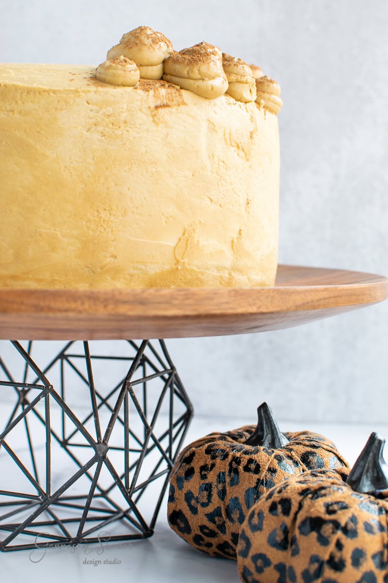 A round yellow cake with decorative dollops on top sits on a wooden cake stand with a modern geometric metal base. Below the stand are two small pumpkins with a leopard print design, adding a farmhouse touch. The background is a light gray.