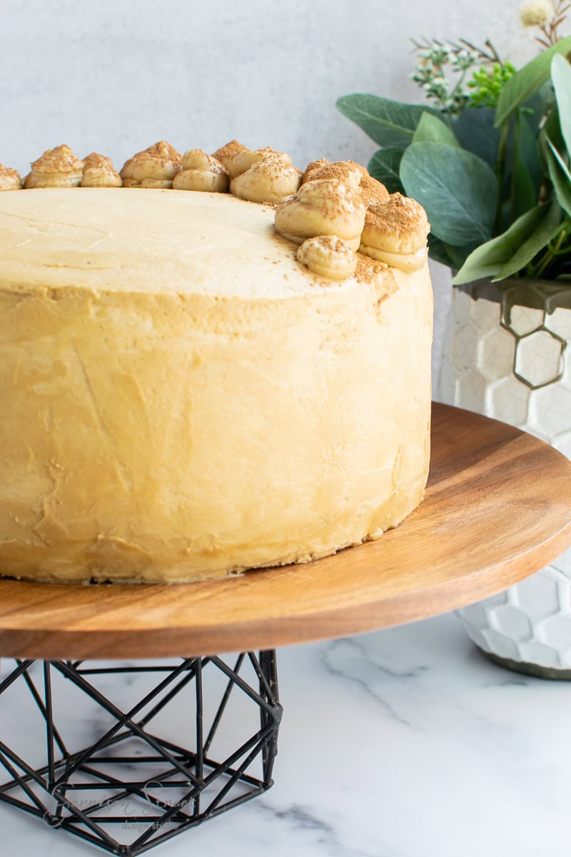 A close-up of a round cake with light brown frosting and decorative dollops on top, elegantly displayed on a farmhouse-style wooden cake stand with a black metal base. A potted plant with green leaves adds a touch of modern charm in the background.