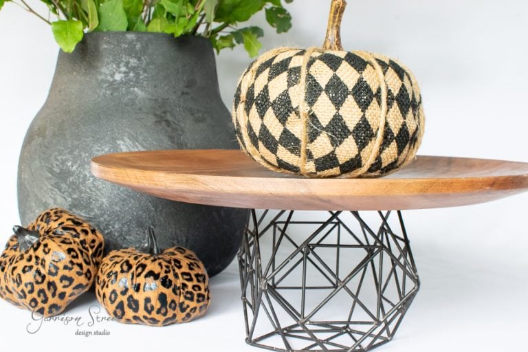 A decorative black and gold harlequin-patterned pumpkin sits elegantly on a wooden cake stand with a geometric metal base. Nearby, two leopard-print pumpkins add flair in front of a large, dark vase filled with greenery, creating a striking farmhouse-modern blend.