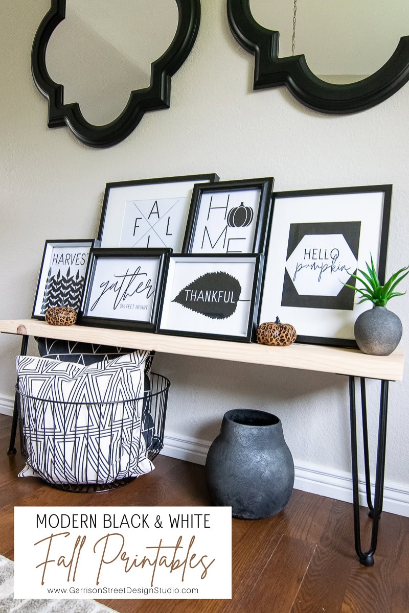 A modern display of black and white fall-themed prints is arranged on a wooden bench with hairpin legs. Below, a geometric-patterned basket and gray vase add style. Two small pumpkins sit on the bench, enhancing the autumn motif with their charming presence, perfect for showcasing seasonal printables.
