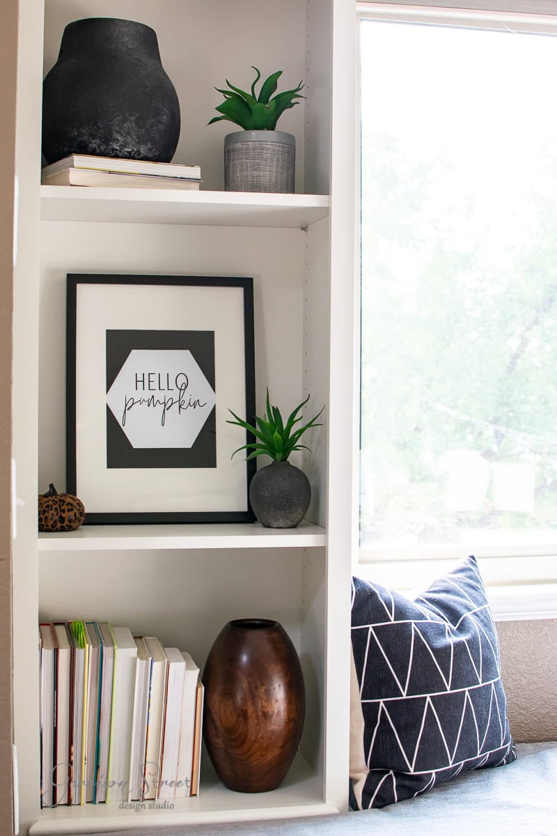 A modern white bookshelf adorned with plants, a framed "Hello Pumpkin" fall print, decorative vases, and colorful books stands elegantly. A cushioned bench with a geometric-patterned pillow sits beside it, next to a window framing views of lush trees.