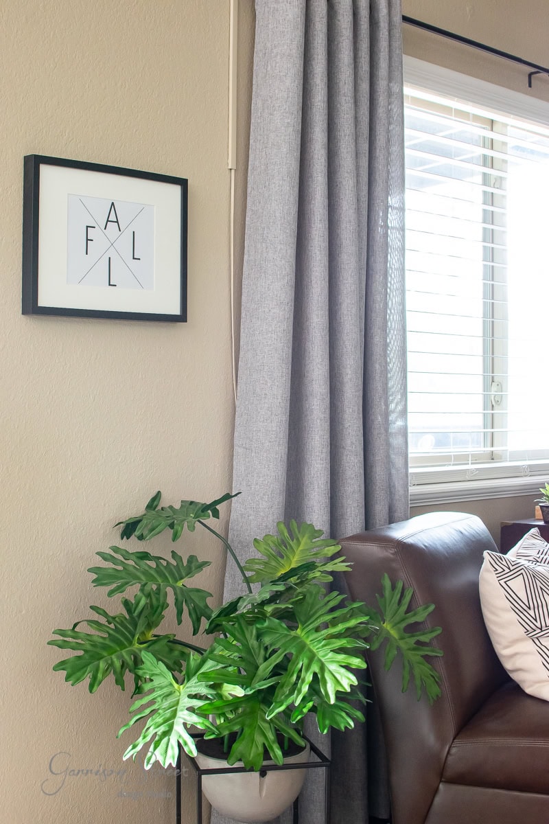 A cozy, modern living room corner featuring a brown leather sofa and a green potted plant. A gray curtain drapes by the window, while framed artwork with the word "FALL" adds charm to the beige wall, reminiscent of stylish printables brought to life.