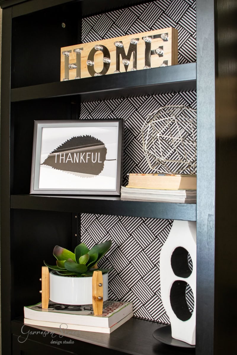 A modern black shelving unit displays decorative items: a "HOME" sign, framed "Thankful" leaf print, geometric sculpture, books, a succulent in a white pot on wooden legs, and a white abstract sculpture. The backing features a black and white checkered pattern that enhances the Fall vibe.
