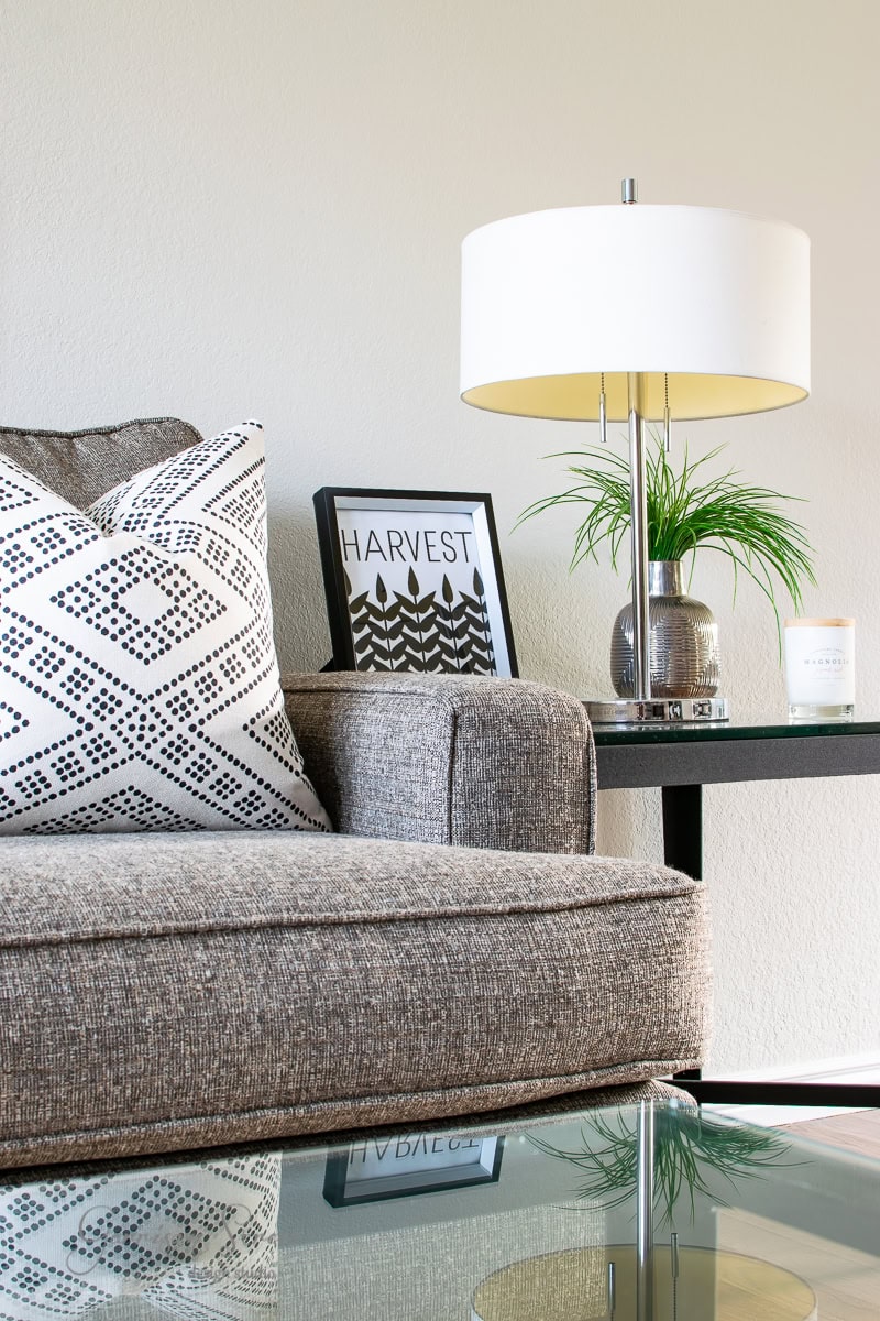 A cozy living room corner showcases a gray sofa adorned with a black and white patterned pillow. A small side table holds a modern lamp, fall-themed printables, a candle, and a vase with a green plant. A glass coffee table is elegantly placed in the foreground.