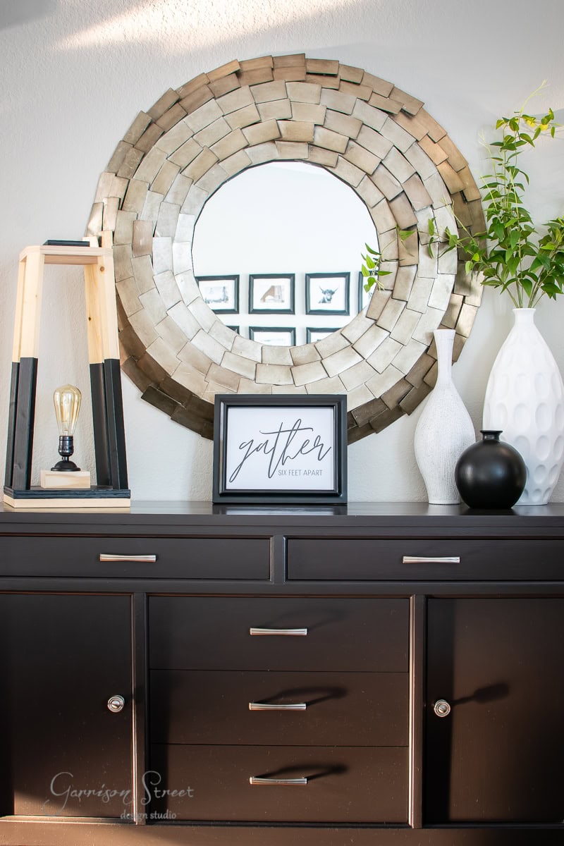 A modern dresser topped with decorative items features a unique layered circular mirror, vases, a framed printable art piece, and a potted plant. A framed sign reads "gather." The setting beautifully blends rustic charm with contemporary elements perfect for fall.