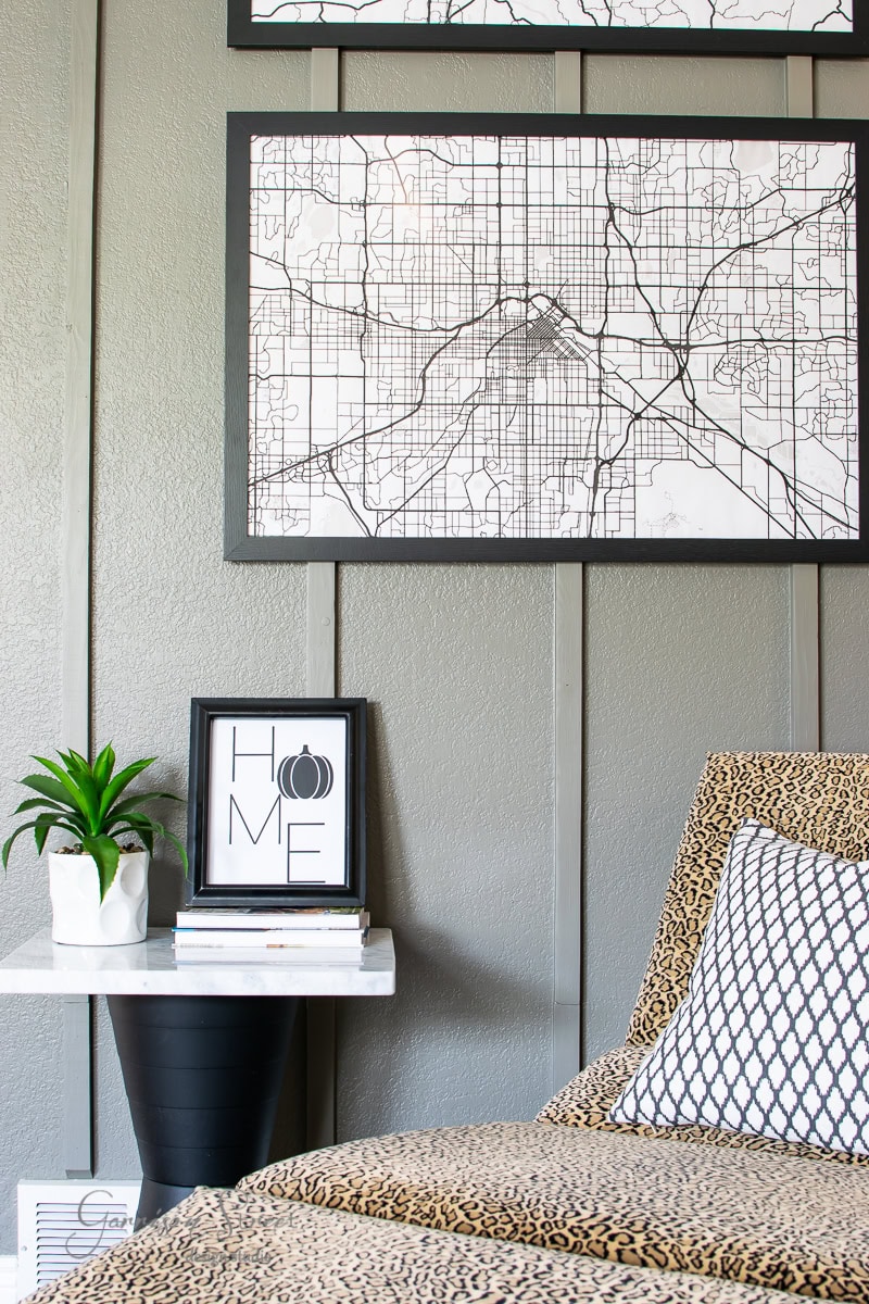 A stylish, modern bedroom interior features a grayscale map on the wall and a white side table holding fall-inspired potted plants, books, and framed printables with "HOME" text. The bed is adorned with patterned cushions and an animal print blanket.