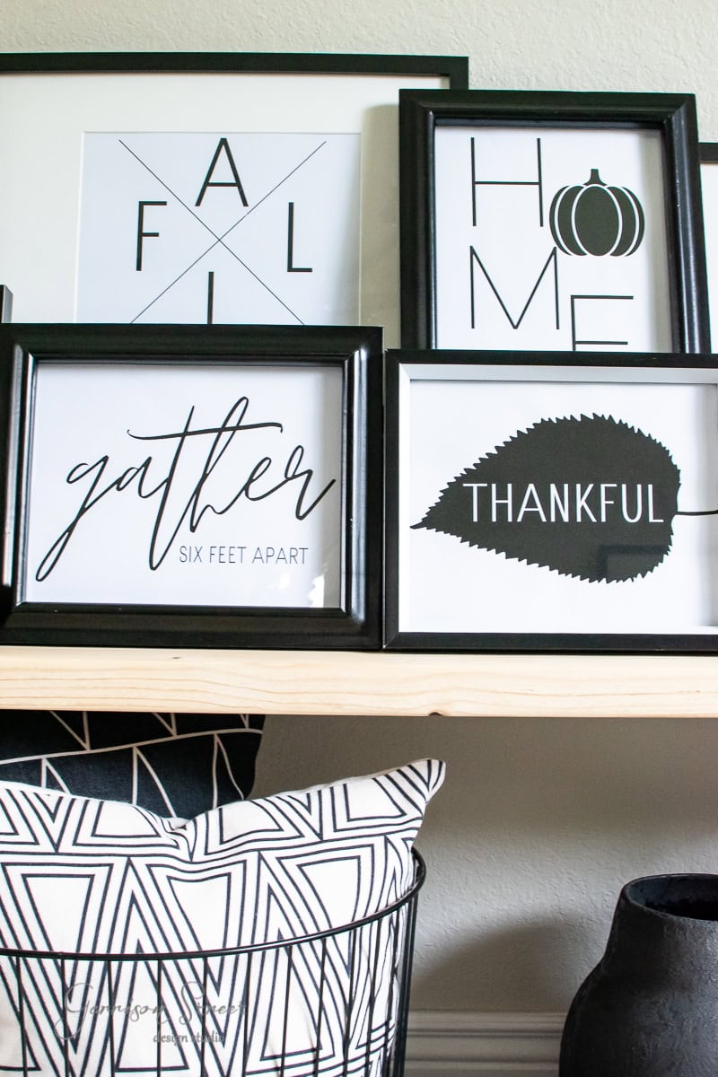 A set of modern, framed autumn-themed prints adorns a shelf. One reads "GATHER SIX FEET APART," another says "THANKFUL" with a leaf design. There's also prints with the words "FALL" and "HOME" featuring pumpkin silhouettes. A patterned pillow is nestled below.