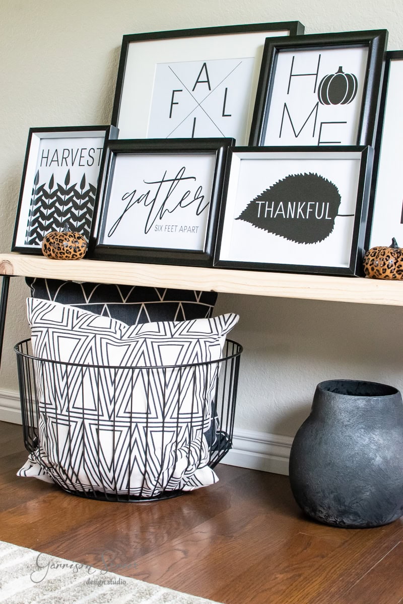 A modern decorative display on a wooden shelf features framed fall-themed printables with stylish designs. Below, a wire basket holds geometric-patterned pillows, and a sleek black vase sits on the floor. Small pumpkin decorations accent the scene.