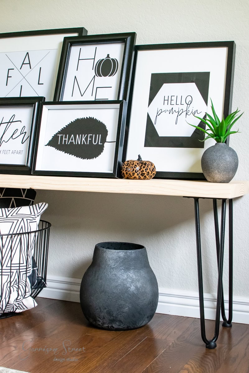 A decorative shelf displays framed art with words like "Thankful" and "Hello Pumpkin." Embrace the modern Fall vibe with a small pumpkin figurine and potted plant. Below, a wire basket holds pillows beside a large black vase. Add seasonal printables for a personalized touch.
