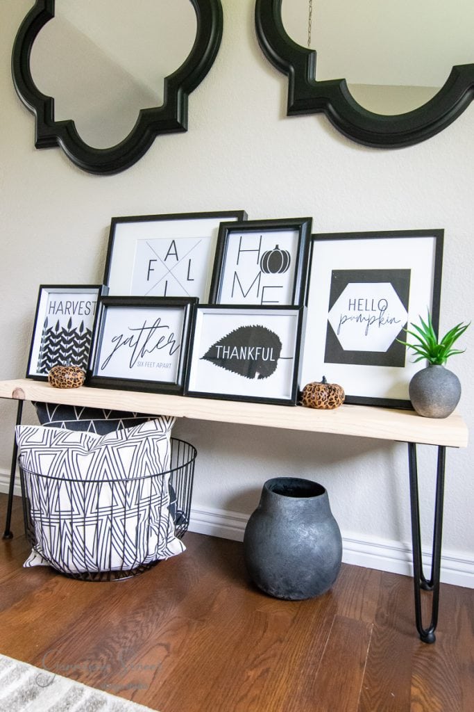 Free Printables A wooden bench holds five framed motivational prints, including fall-themed words like "gather" and "thankful." Two small plants and decorative bowls adorn the bench. Below, a modern black-and-white patterned basket rests on wooden flooring. Mirrors hang above the bench.
