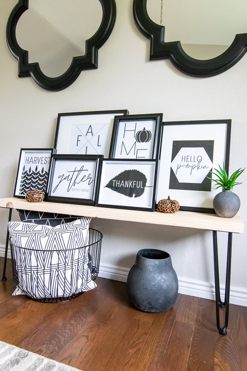 A wooden bench holds five framed motivational prints, including fall-themed words like "gather" and "thankful." Two small plants and decorative bowls adorn the bench. Below, a modern black-and-white patterned basket rests on wooden flooring. Mirrors hang above the bench.