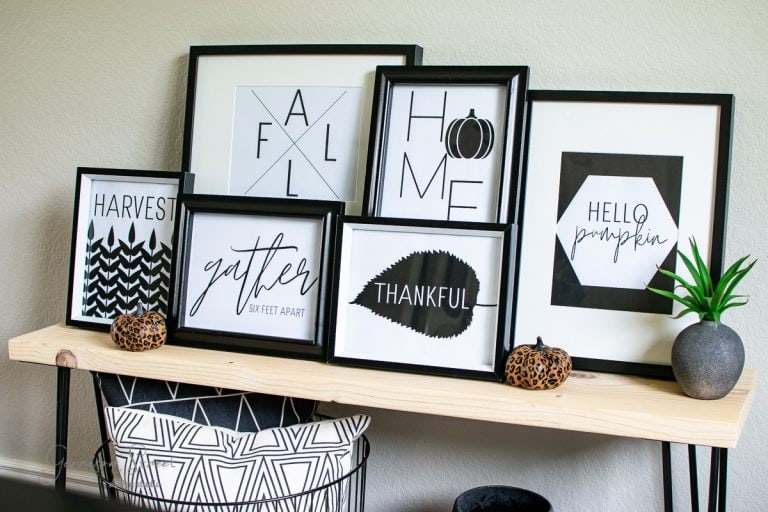 A wooden table showcases seven modern framed prints with fall-themed text and graphics, featuring words like "HARVEST," "gather," "THANKFUL," and "HOME." Two small decorative pumpkins and a potted plant accent the arrangement, perfect for seasonal printables.