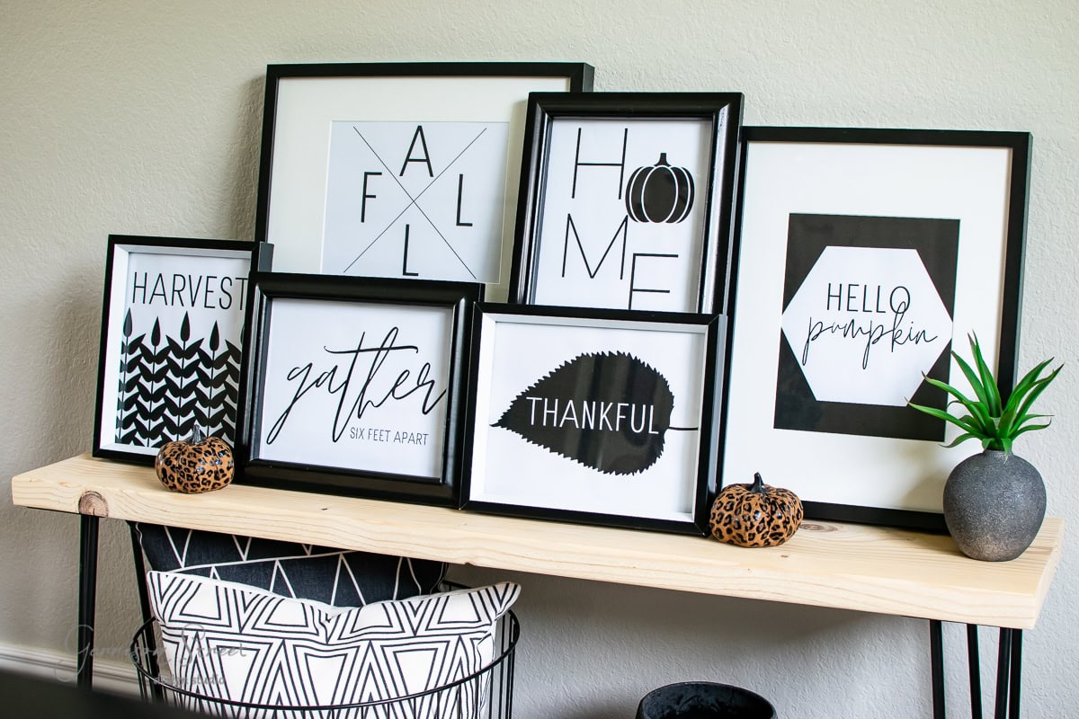 A wooden table showcases seven modern framed prints with fall-themed text and graphics, featuring words like "HARVEST," "gather," "THANKFUL," and "HOME." Two small decorative pumpkins and a potted plant accent the arrangement, perfect for seasonal printables.