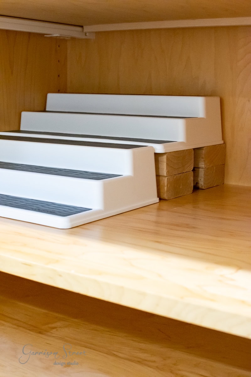 White tiered shelf risers on a light wood shelf with wooden blocks placed underneath for extra height, perfect for efficient pantry organization.