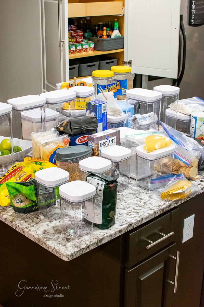 A kitchen counter covered with clear storage containers and various pantry items, such as instant oatmeal, pasta, crackers, and snacks, showcases perfect pantry organization. In the background, cabinets filled with more neatly arranged items line the shelves.