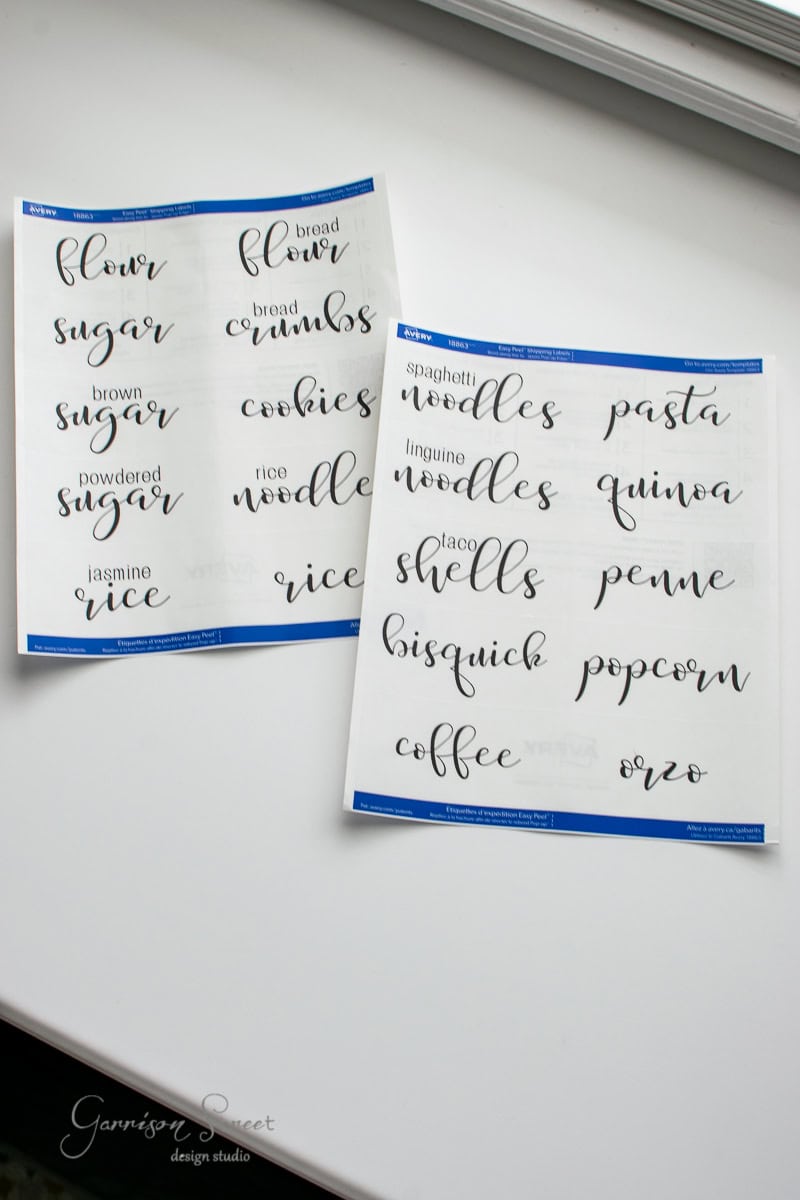 Two white sheets on a table showcase elegant black cursive labels for pantry organization, listing items like flour, sugar, noodles, pasta, rice, quinoa, and coffee. Perfect for a tidy cabinet!.