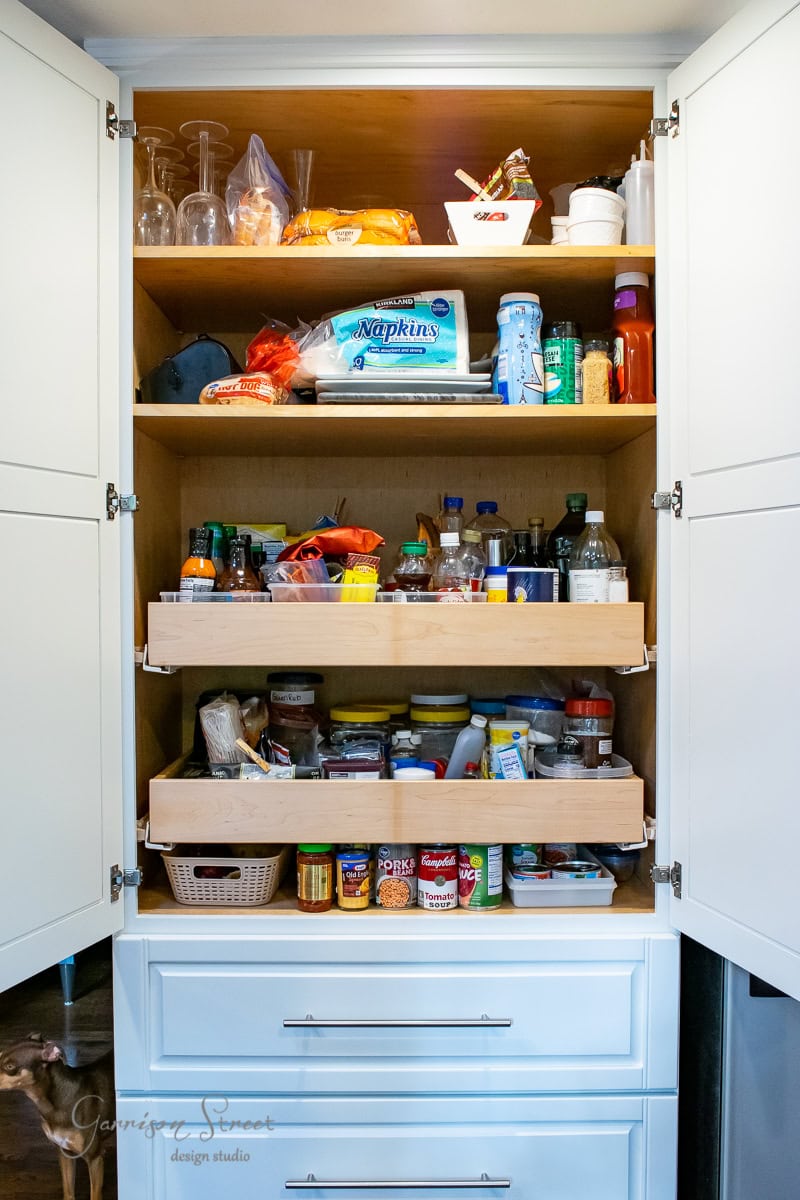 A neatly organized pantry cupboard boasts three shelves. The top shelf holds glasses and napkins, the middle is lined with various bottles and jars, while the bottom brims with canned goods and condiments. Pristine white cabinets elegantly surround these well-stocked shelves.