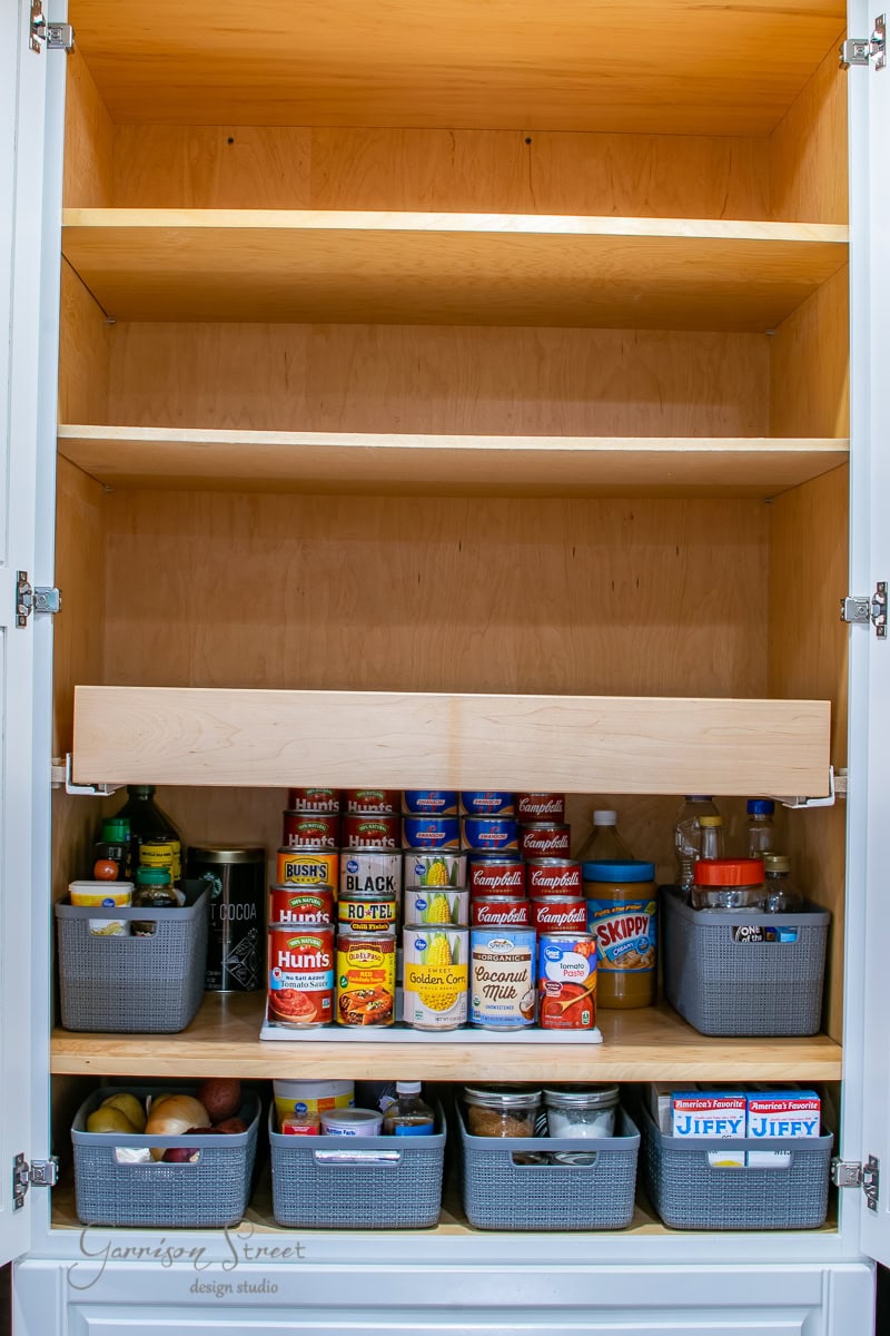 A well-organized pantry with tidy shelves showcases impeccable organization. The top shelves are empty, while the bottom shelf holds canned goods, jars, and bins brimming with various food items. With the door open, it beautifully displays an array of neatly arranged essentials.