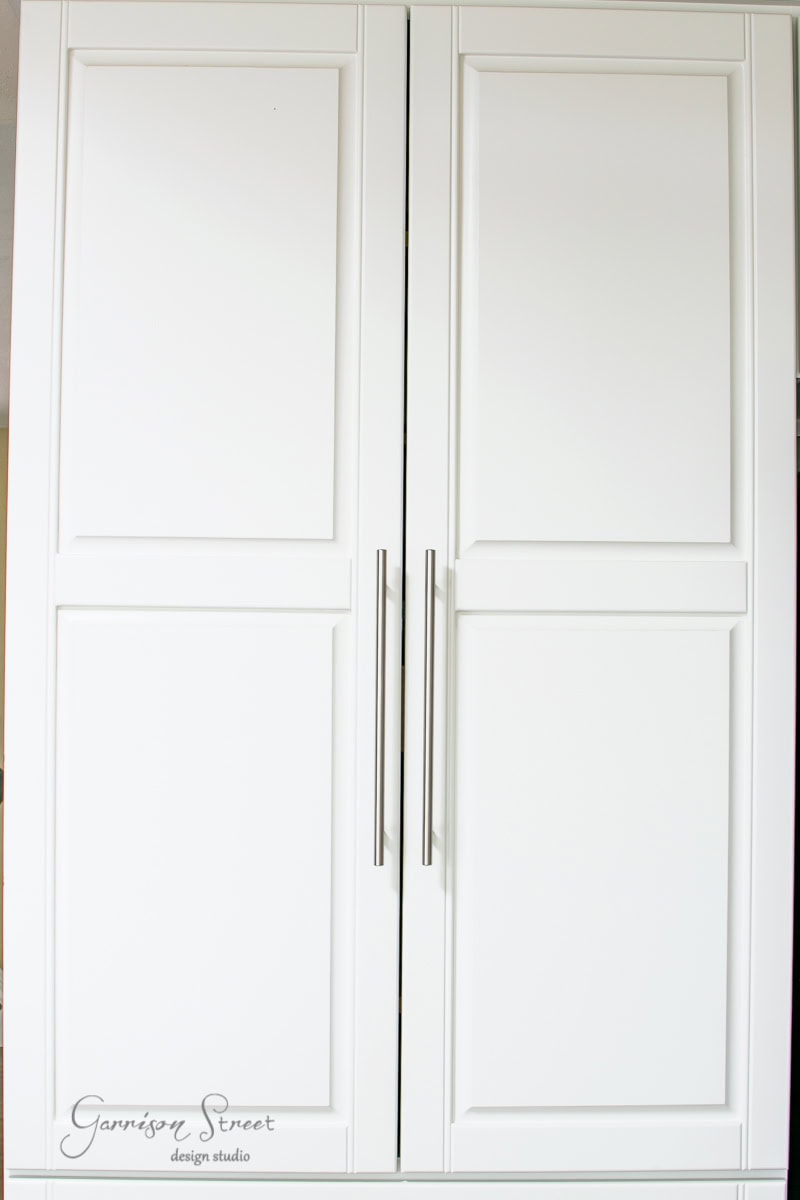 A pair of tall, white cabinet doors with sleek silver handles, offering a minimalist design perfect for organization. The words "Garrison Street design studio" are printed in the lower left corner.