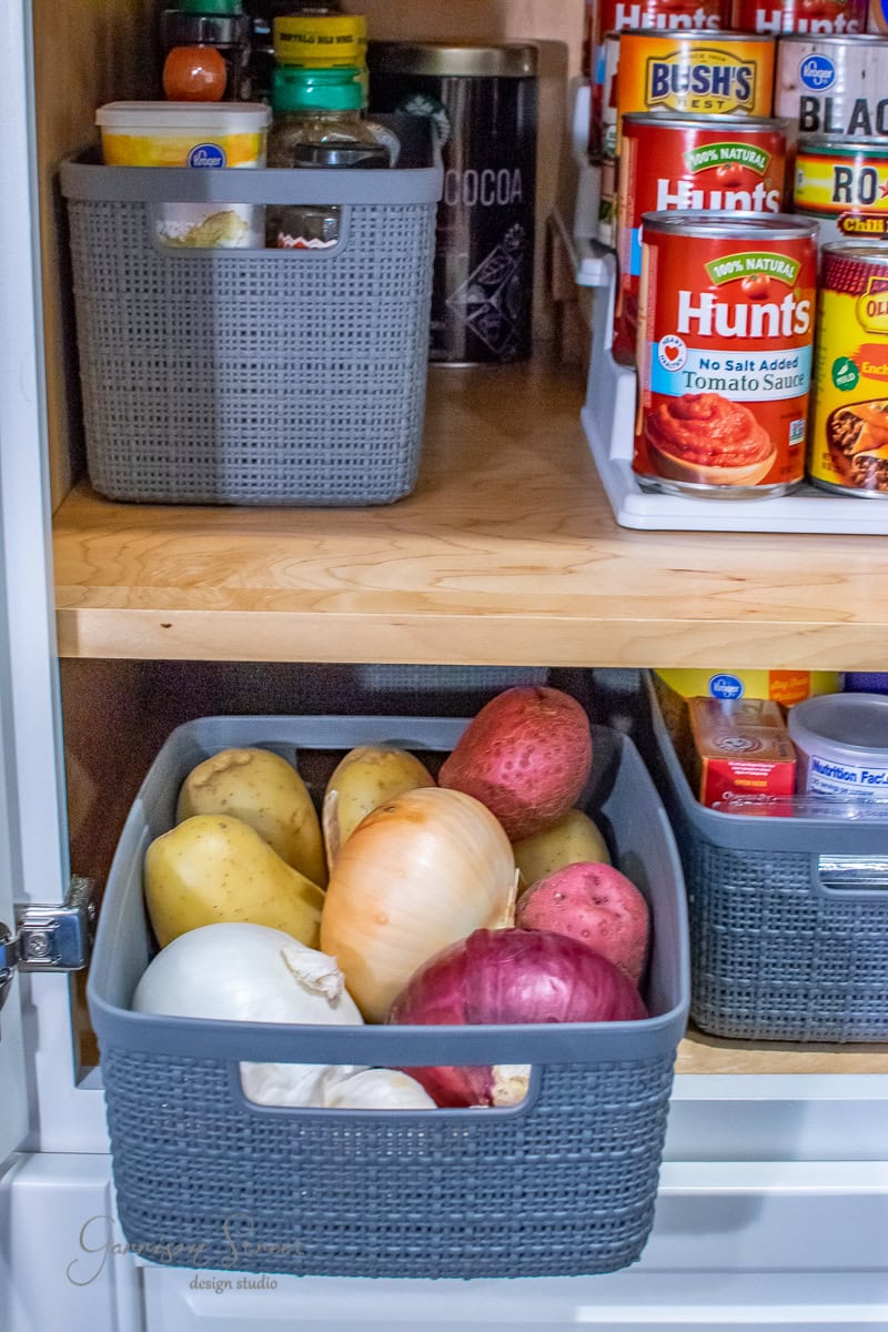 A well-organized pantry showcases gray baskets on wooden shelves. One basket holds assorted onions and potatoes, while another displays small jars. Above, canned goods like beans and tomatoes are neatly stacked in white organizers for optimal cabinet efficiency.