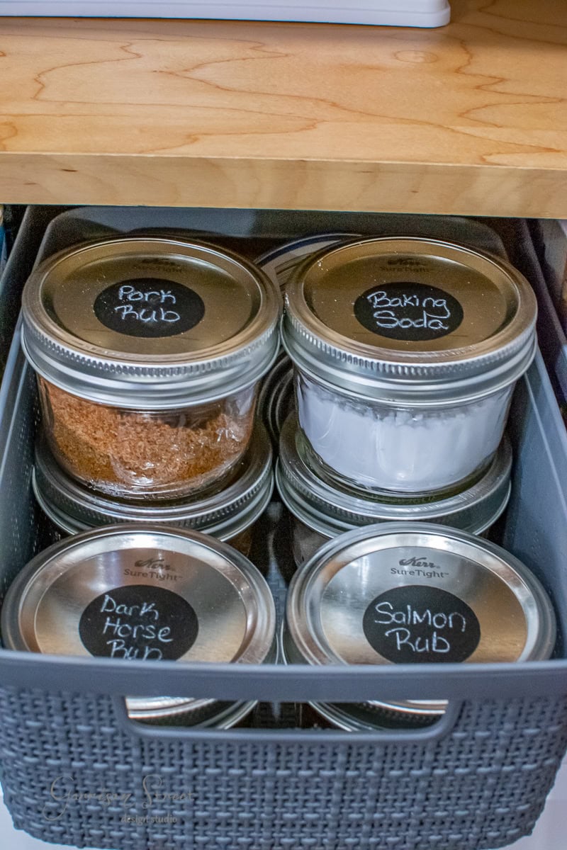 A woven basket sits on a wooden shelf, organizing jars with metal lids. Each jar, labeled with chalkboard stickers, reads: Pork Rub, Baking Soda, Dark Horse Rub, and Salmon Rub. This pantry essential keeps your spices neatly in place.