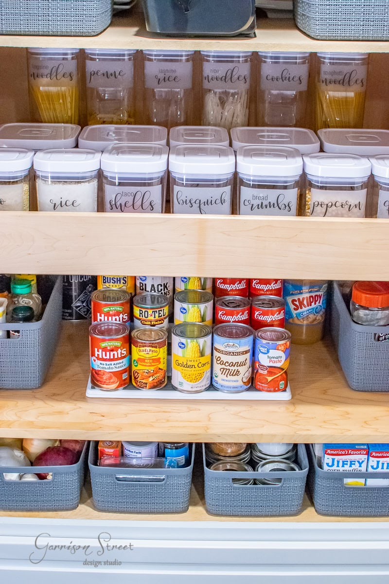 A neatly organized pantry showcases a cabinet with labeled containers for pasta, rice, and snacks on the top shelf. Canned goods and jars, featuring beans and sauces, are arranged on the middle shelf. More containers are stored below in gray bins for optimal organization.