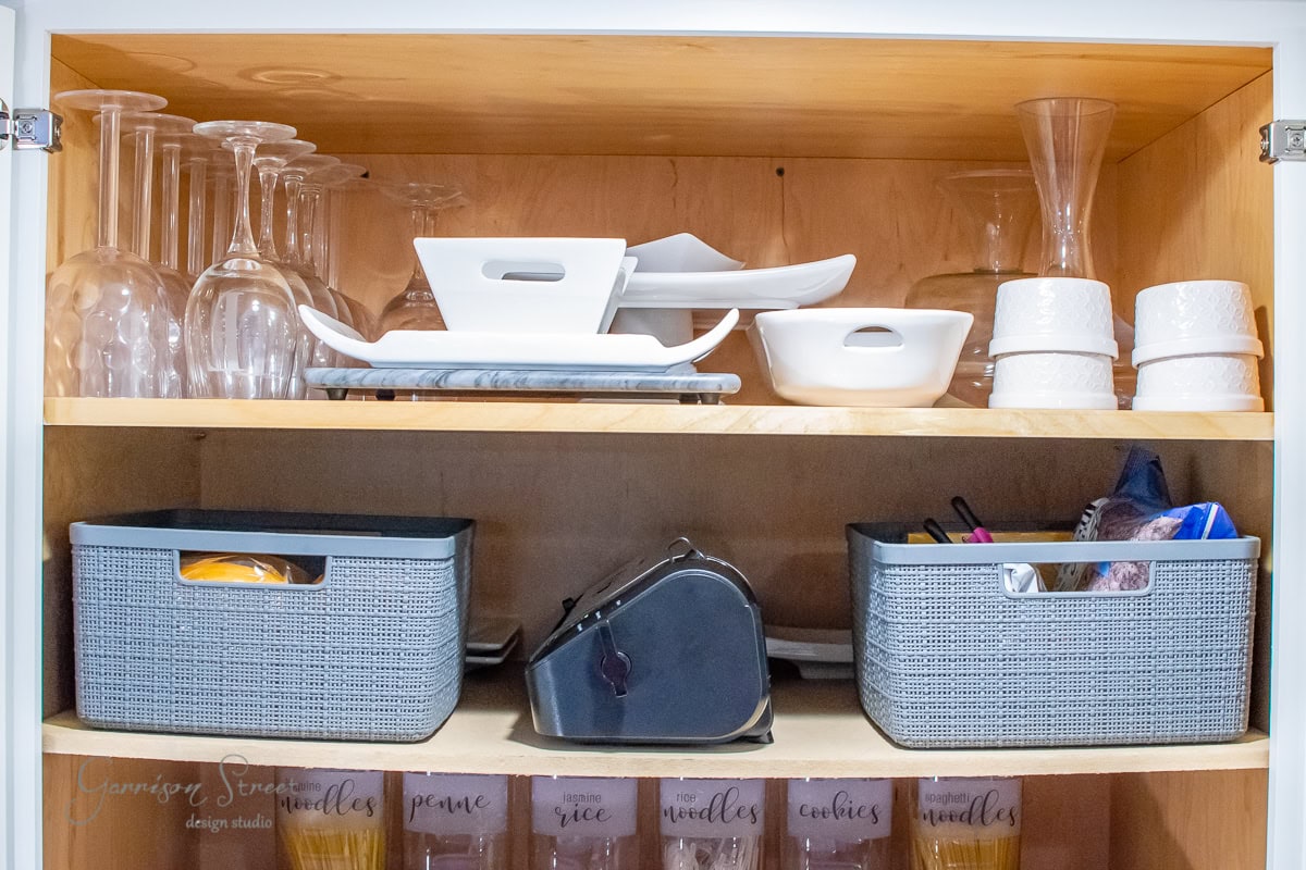 A wooden cabinet showcases wine glasses, white dishes, and bowls on the top shelf. Below are gray storage baskets perfect for pantry organization alongside a label maker. The bottom shelf neatly holds labeled containers filled with grains and pasta.