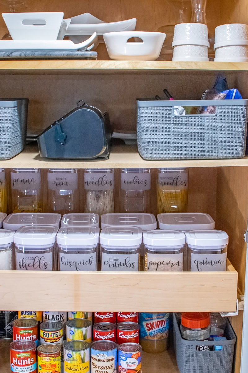 A neatly organized pantry features labeled containers. The shelves, a testament to perfect organization, hold baskets with dishes, glass jars, and containers labeled with food items like pasta, bread crumbs, and quinoa. Canned goods and jars are stored efficiently on the lower shelves.