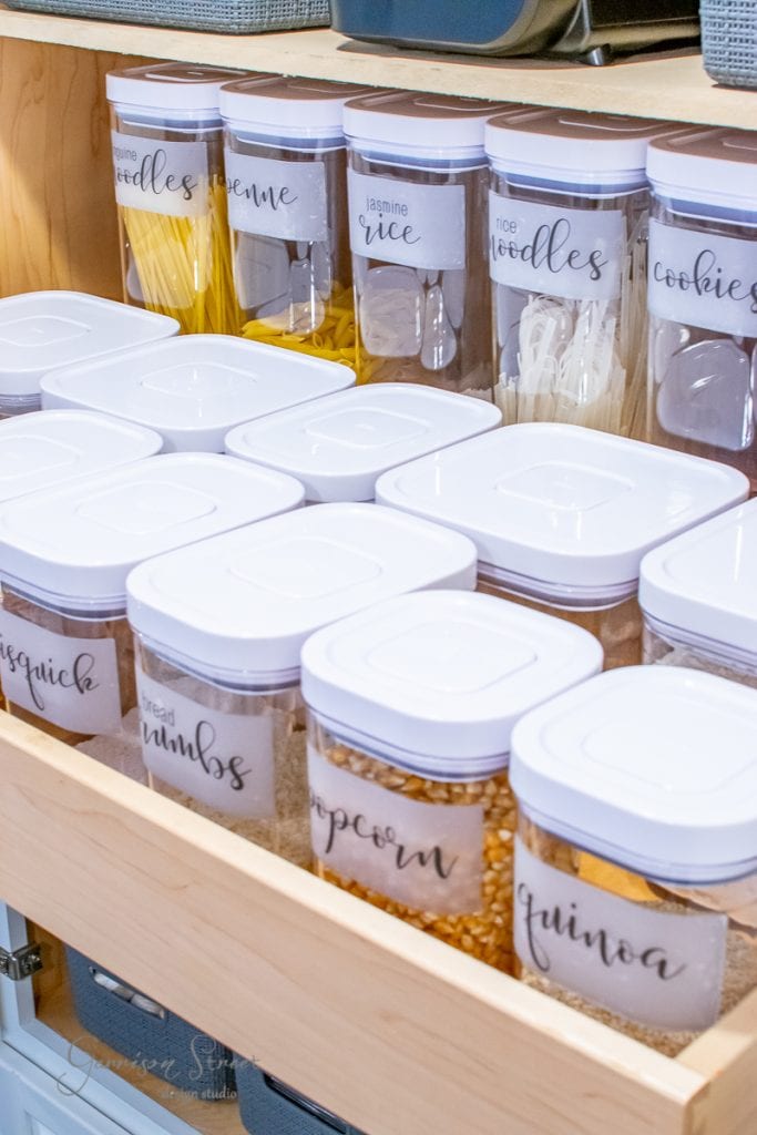 Free Printables A kitchen pantry drawer filled with neatly organized, labeled containers. Visible labels include noodles, rice, cookies, popcorn, breadcrumbs, and quinoa. The transparent containers with white lids showcase a tidy array of dry food items.