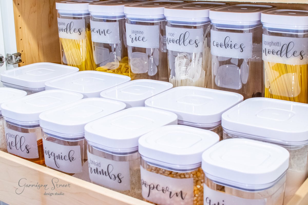 A pantry shelf with clear plastic containers of various dry goods, labeled for items like rice, noodles, cookies, and popcorn. The containers have white lids and are neatly organized in rows.