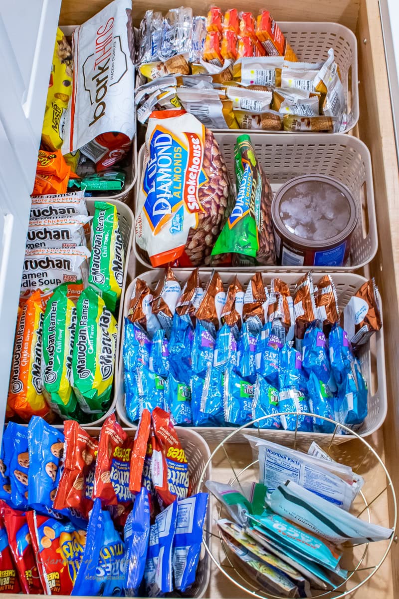 A drawer organized with various snacks, including ramen noodle packs, granola bars, almonds, crackers, and fruit snacks. Items are neatly arranged in baskets for easy access and separation, adding a touch of pantry organization.