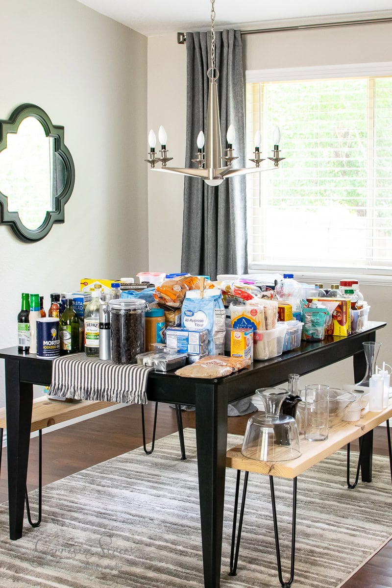 A dining room showcases a black table adorned with snacks, beverages, and pantry staples, neatly organized under a striped cloth. Benches flank either side while a modern chandelier illuminates the scene, creating an ideal space for culinary inspiration beyond the nearby cabinet.