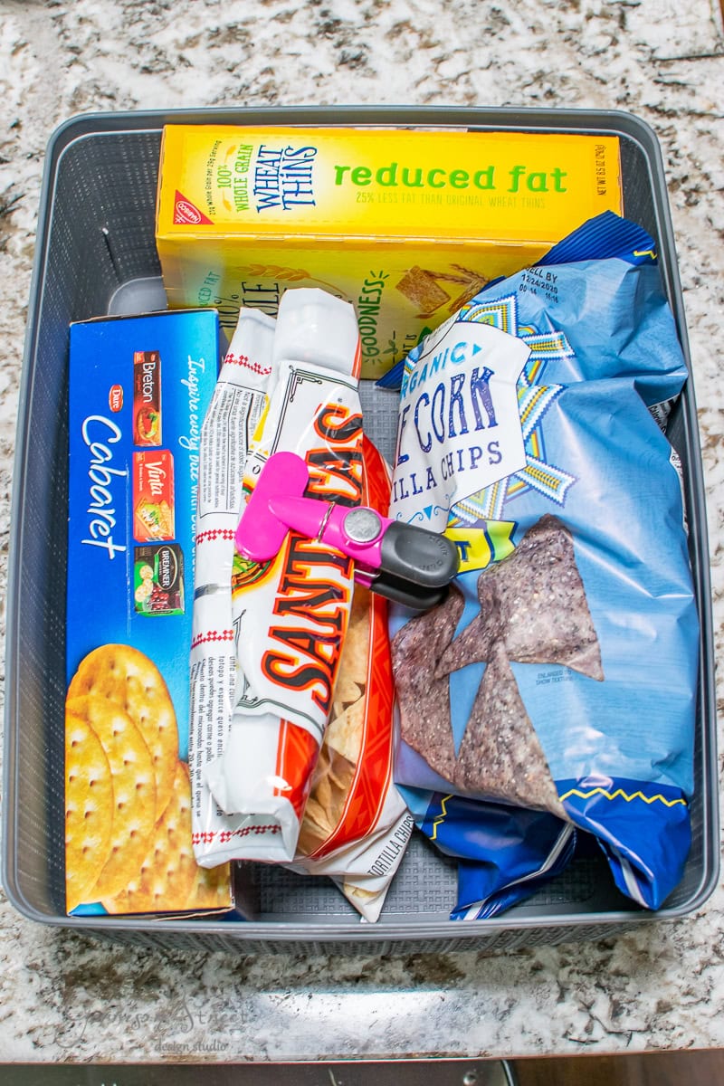 A storage basket filled with various snack items, including reduced-fat wheat crackers, Saltines, organic blue corn tortilla chips, and Goya cookies, is neatly organized on a speckled countertop, ready to enhance your pantry cabinet's appeal.