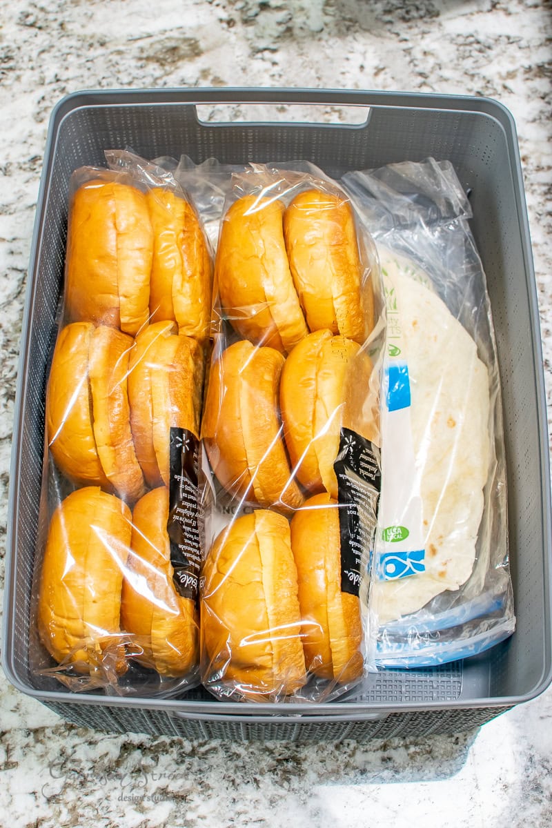 A gray basket containing packages of burger buns and tortillas, all wrapped in plastic, sits neatly on the granite countertop, showcasing a touch of pantry organization.
