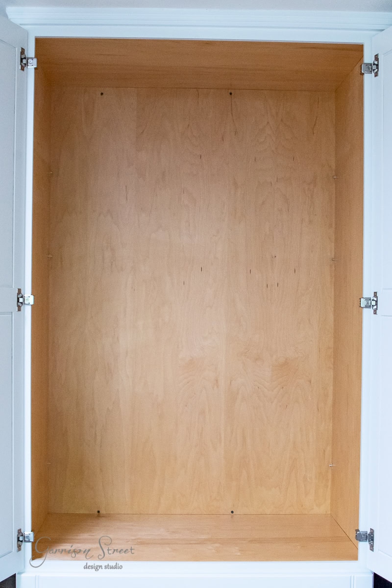 An empty wooden pantry cabinet with open white doors and silver hinges displays a smooth, light-colored wood interior. The tall, spacious design offers ample organization opportunities with its simple and clean lines.
