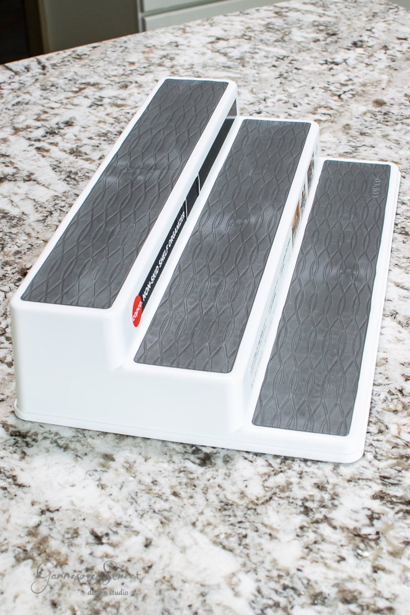 A multi-tiered spice rack sits on a speckled granite countertop. The white rack, with three gray, non-slip steps, is perfect for pantry organization and designed to display spices or similar items.