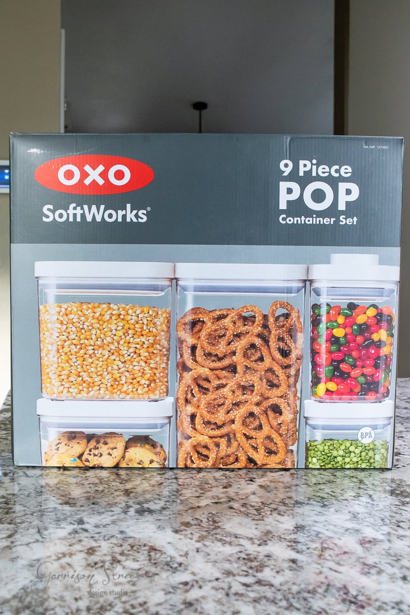 A box of OXO SoftWorks 9 Piece POP Container Set is displayed on a kitchen countertop, enhancing pantry organization. The packaging showcases various dry foods like popcorn, pretzels, and candies neatly stored in clear containers.