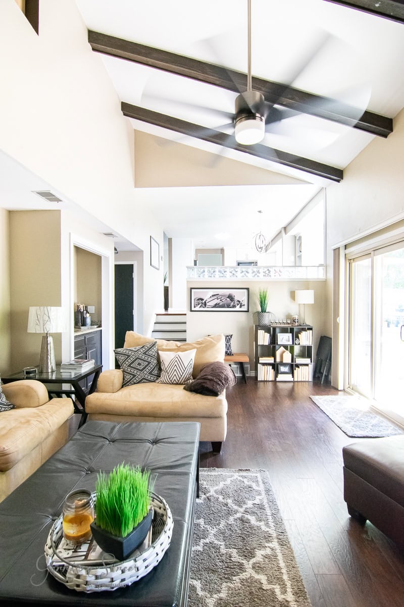 A modern living room with high, beamed ceilings and wooden floors boasts a casual elegance. It includes tan leather sofas, a patterned rug, and a black ottoman with decor. A ceiling fan spins above as natural light streams through the sliding glass door. Shelving and art complete the makeover.