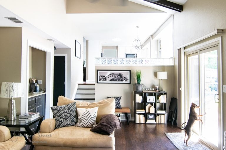 A modern family room features a beige sofa adorned with decorative pillows and a sleek dark wooden floor. A small dog stands by the glass door on the right, while artwork and a plant enhance the shelves near the staircase, completing this stylish makeover.