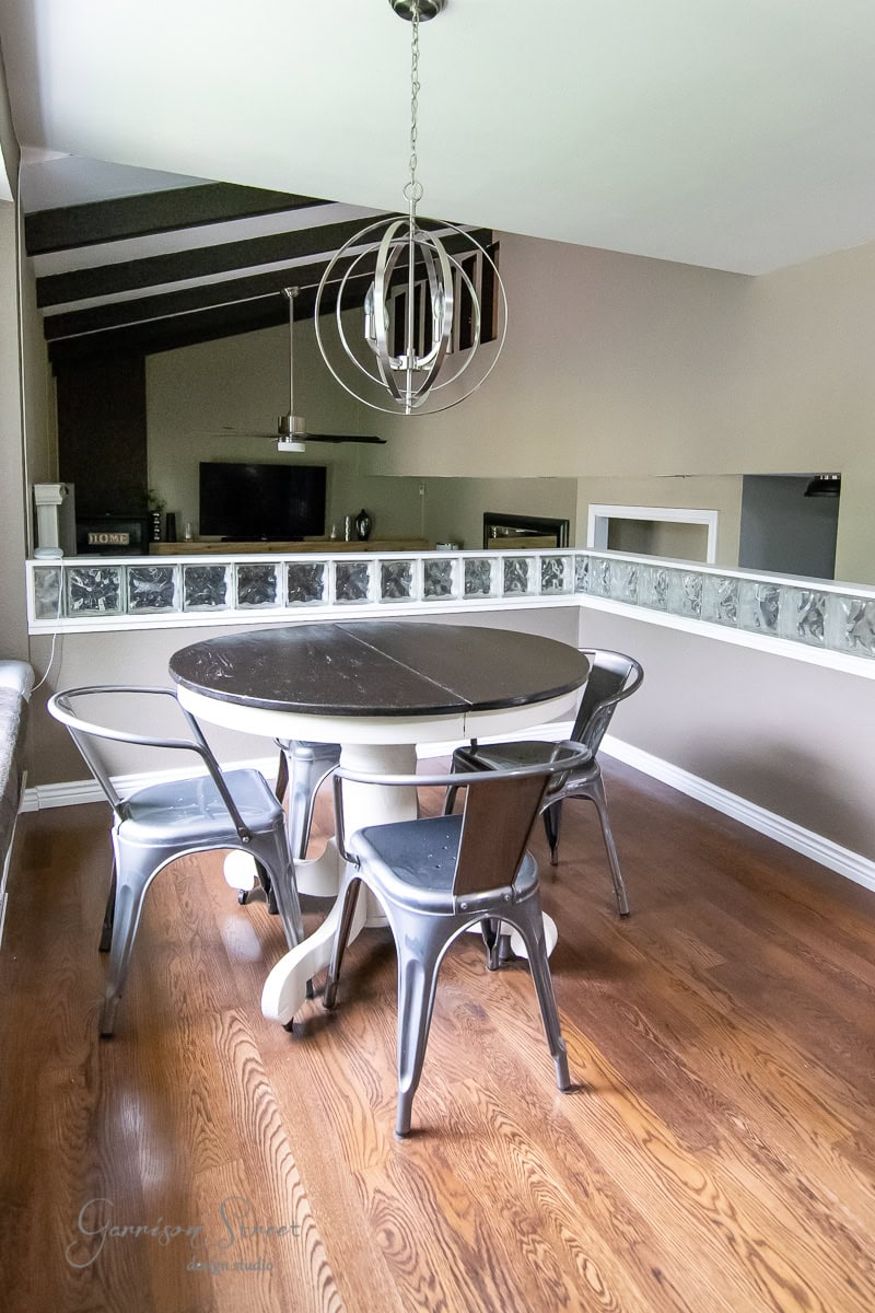 A modern dining area features a round table with four metal chairs on a wooden floor. Above, a spherical pendant light, suspended by sleek cables, adds a contemporary touch. The wall is partially glass-blocked, letting additional light into the space.