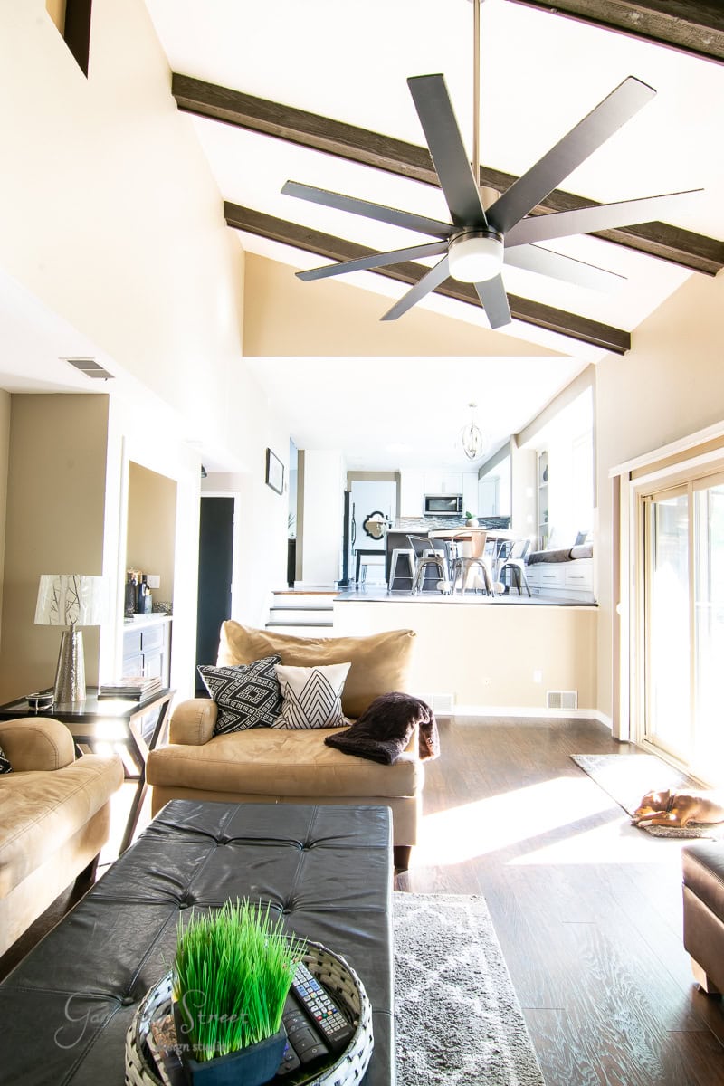 Spacious living area with high ceilings and exposed beams, featuring a large ceiling fan. The room includes a brown sofa, decorative pillows, and a table with a plant. Sunlight streams in through sliding doors, highlighting the view of the dining area on a raised platform with sleek cable railing.