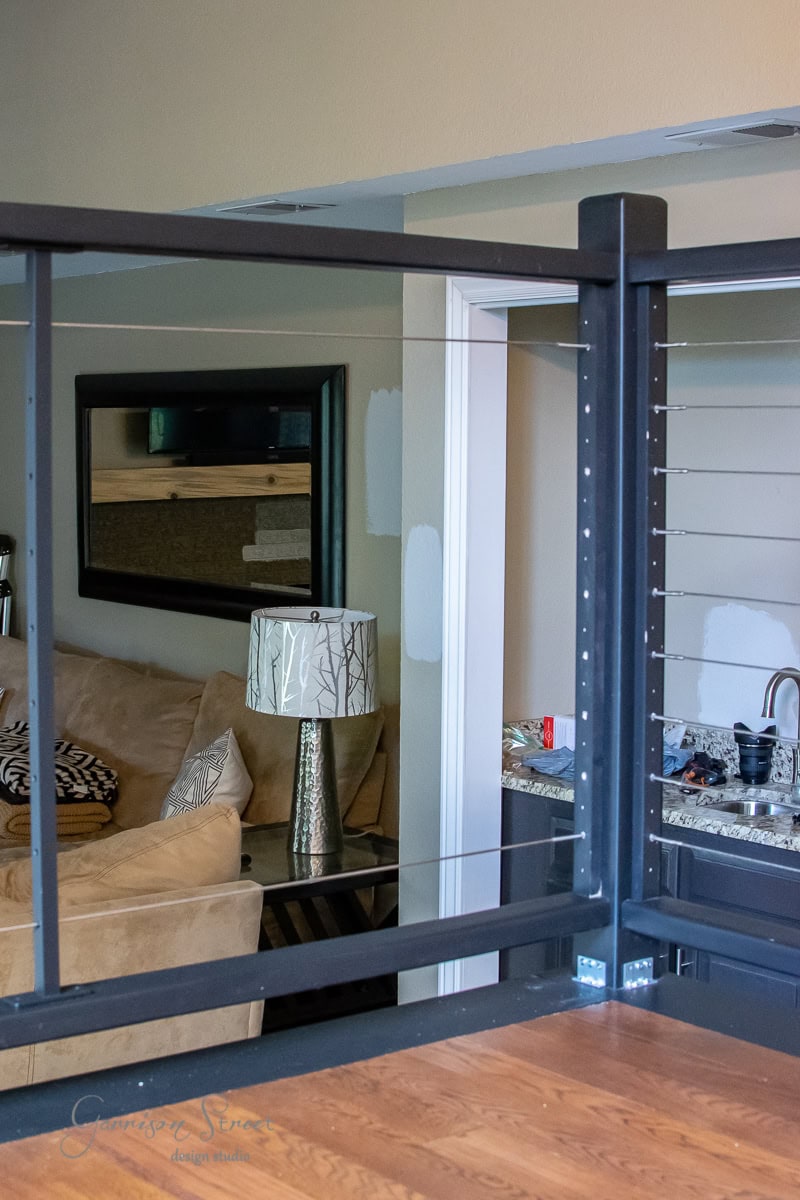 A modern living room features a beige sectional sofa, decorative pillows, and a tree-patterned lamp. A mirror reflects the DIY touch with sample paint patches on the wall. In the foreground, a sleek cable railing adds contemporary flair next to the countertop with a sink in view.