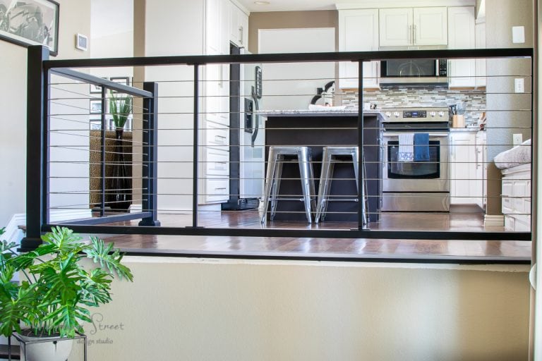 A modern kitchen with white cabinets, stainless steel appliances, and a central island featuring two sleek metal stools. The space is elegantly defined by a black cable railing that separates the kitchen from the lower level, while a small green plant adds a touch of nature in the foreground.