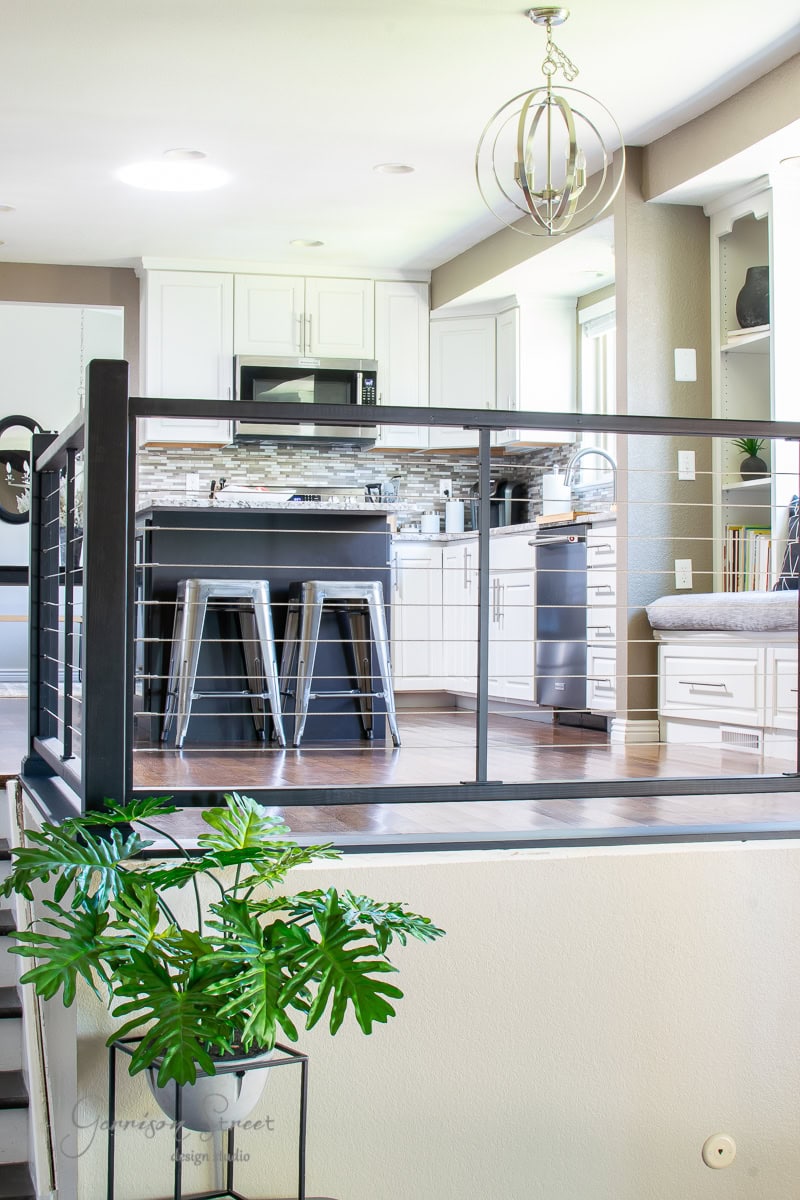A modern kitchen features white cabinets and a black island, complemented by three metallic barstools. A sleek hanging light fixture illuminates the space, while a glass railing separates it from the lower level. A potted plant adds a touch of nature to this elegant DIY-inspired design.