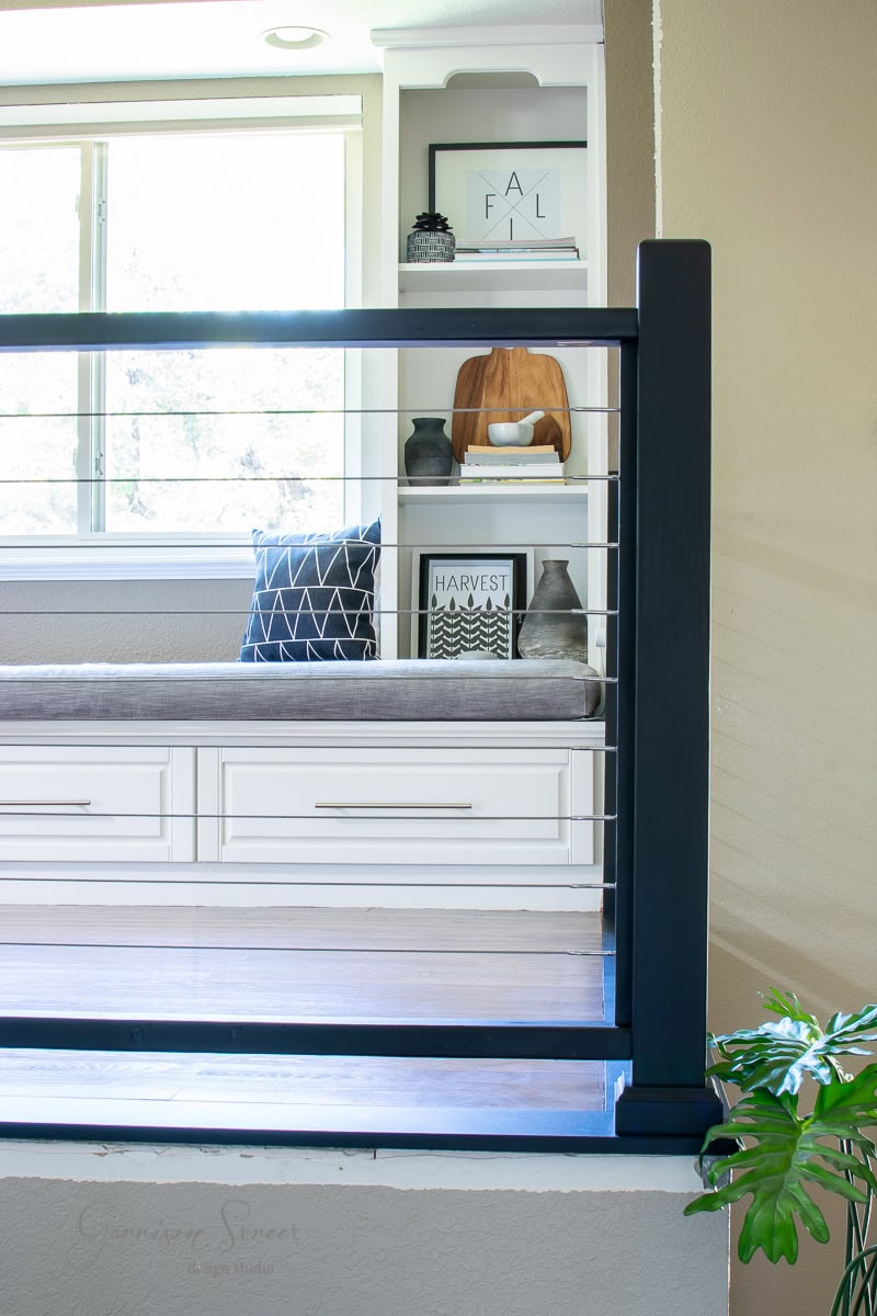 A cozy corner features a white built-in bench with patterned cushions and a throw pillow. Behind it are shelves showcasing decorative items, including a framed "HARVEST" sign, a small plant, and a cutting board. A sleek DIY black cable railing is in the foreground.