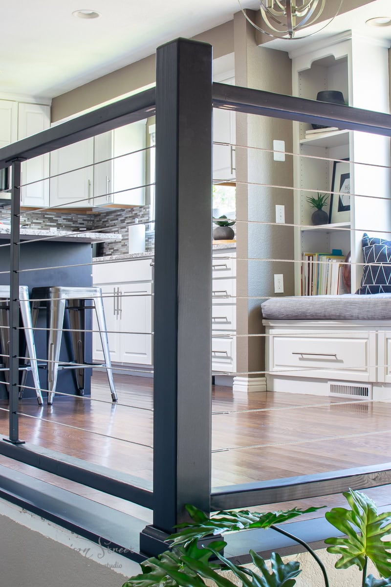 Modern kitchen with white cabinets, a tiled backsplash, and wooden floors. A black cable railing divides the kitchen and adjacent room. The space features built-in shelves and a cozy DIY window seat with cushions, while potted plants add a touch of nature to the foreground.