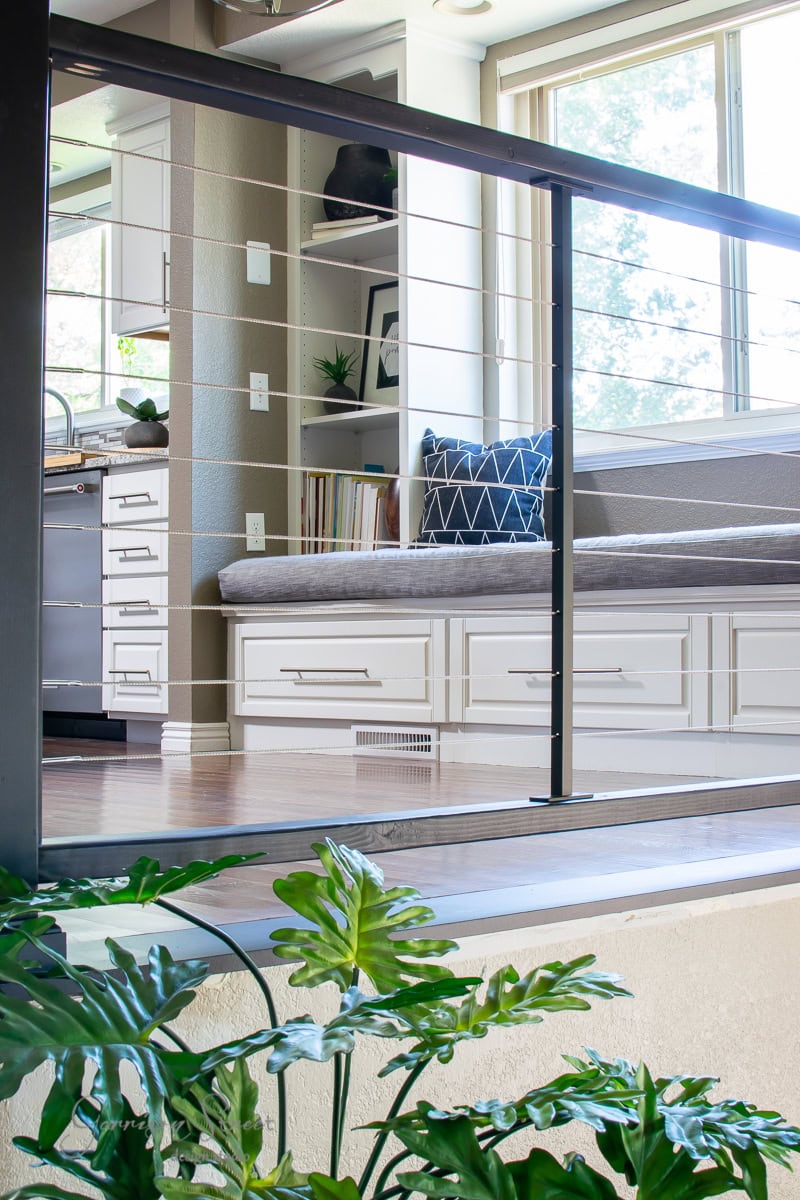 Modern interior with a cozy reading nook featuring a cushioned bench and decorative pillow, accented by a DIY touch. The nook is adjacent to a kitchen area with white cabinets and a sunlit window above, while green plants thrive beside sleek cable railings in the foreground.