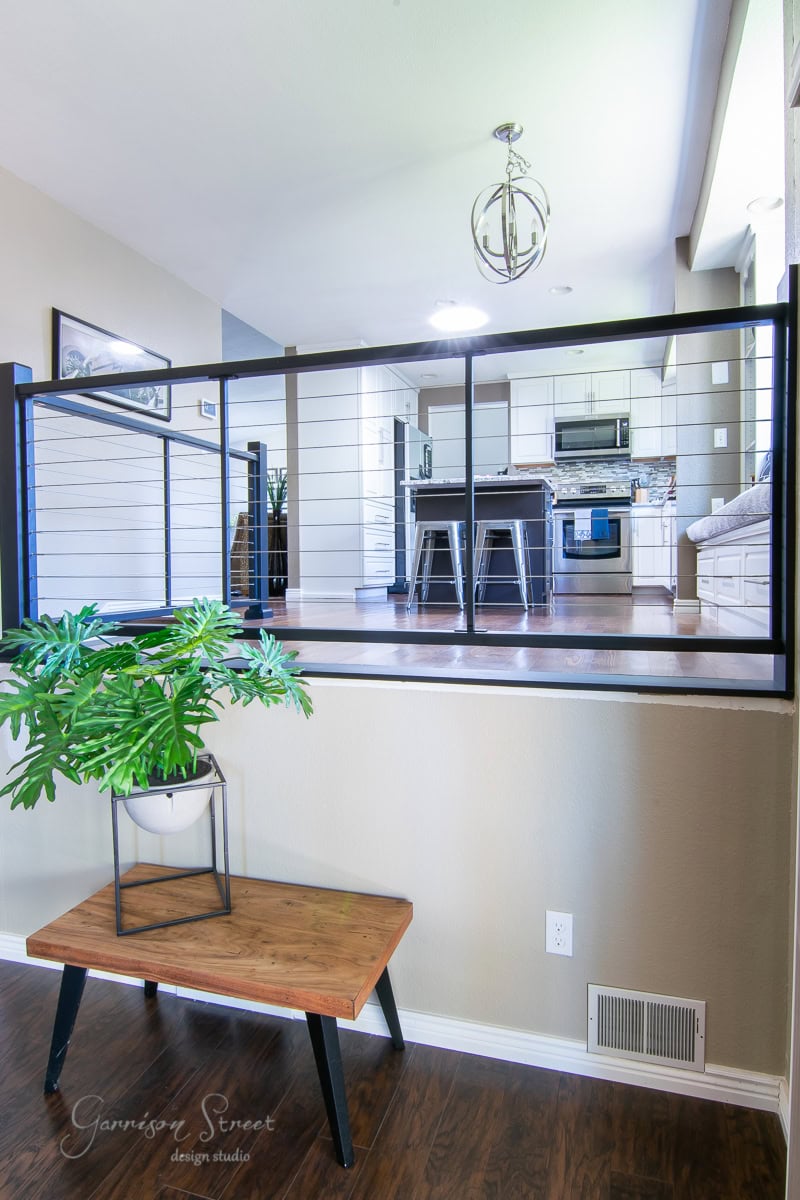A modern home interior featuring a kitchen with stainless steel appliances and a breakfast bar with stool seating. A sleek black railing divides the space, providing a stylish touch. The lower level boasts a wooden bench and a potted plant, perfect for your next DIY project.